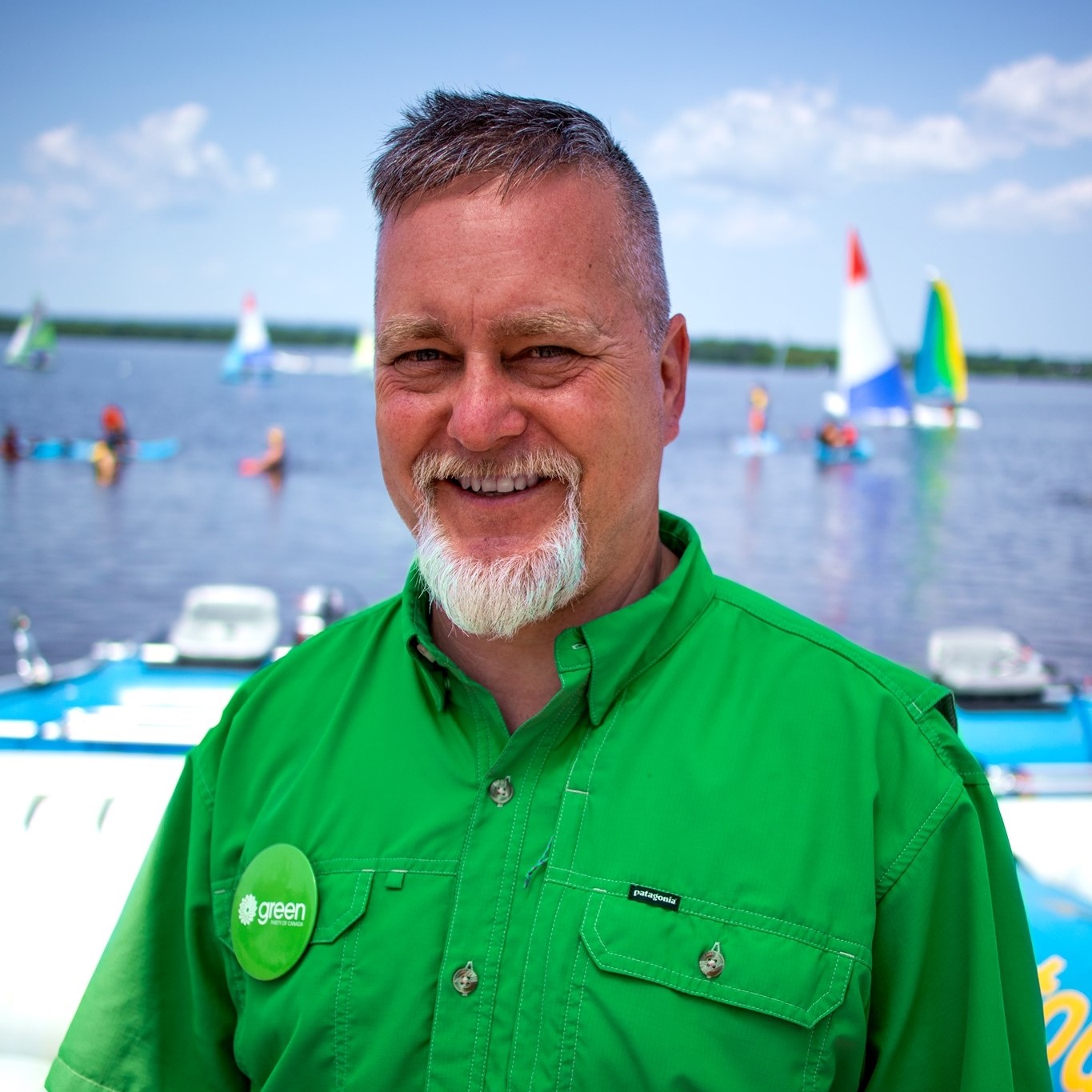 A man in a green shirt and with a grey goatie stands in front of a lake. There is a sail boat on the lake.